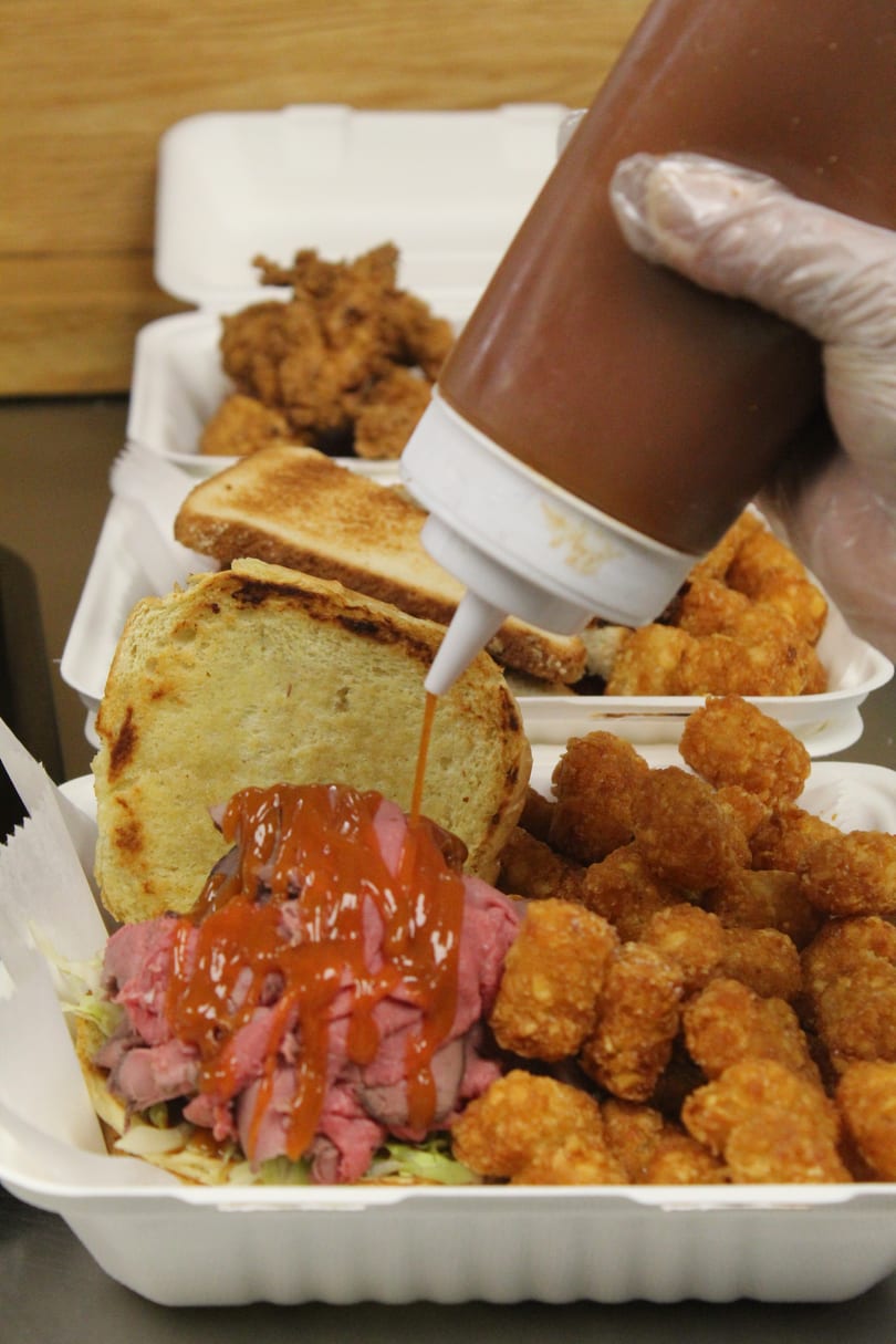 Gloved hand squeezing sauce onto a roast beef sandwich with tater tots, fried chicken, and toast in takeout containers.