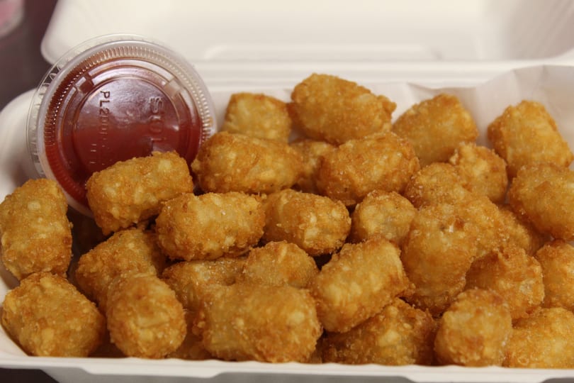 Crispy tater tots and a small cup of ketchup in a white takeout box.